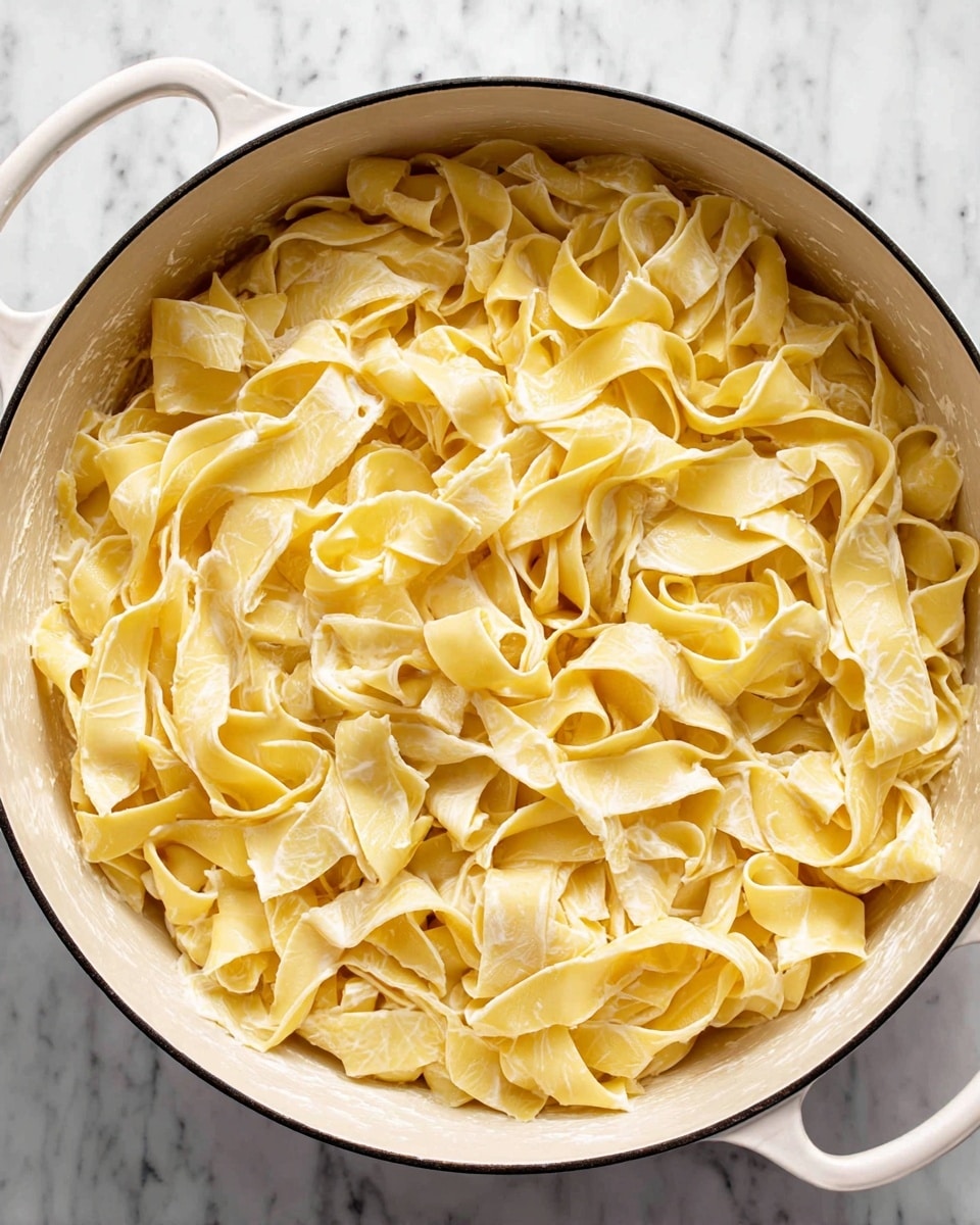 A large white pot filled with wide, flat pasta ribbons piled closely together in layers. The pasta is light yellow with a smooth, creamy white sauce coating each piece, creating a glossy texture with soft folds and curls throughout. The pot is set on a white marbled surface. photo taken with an iphone --ar 4:5 --v 7