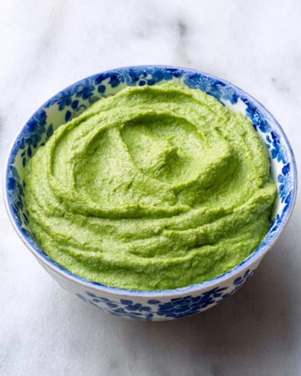 A close-up image of a blue and white patterned bowl filled with thick, smooth green dip. The dip has a creamy texture with slight lumps. Around the bowl are many pale yellow triangular chips with a slightly rough texture. A woman's hand with shiny light-colored nail polish is holding one chip, dipping it into the green dip. The background has a soft blurred blue cloth and the scene is set on a white marbled surface. Photo taken with an iphone --ar 4:5 --v 7