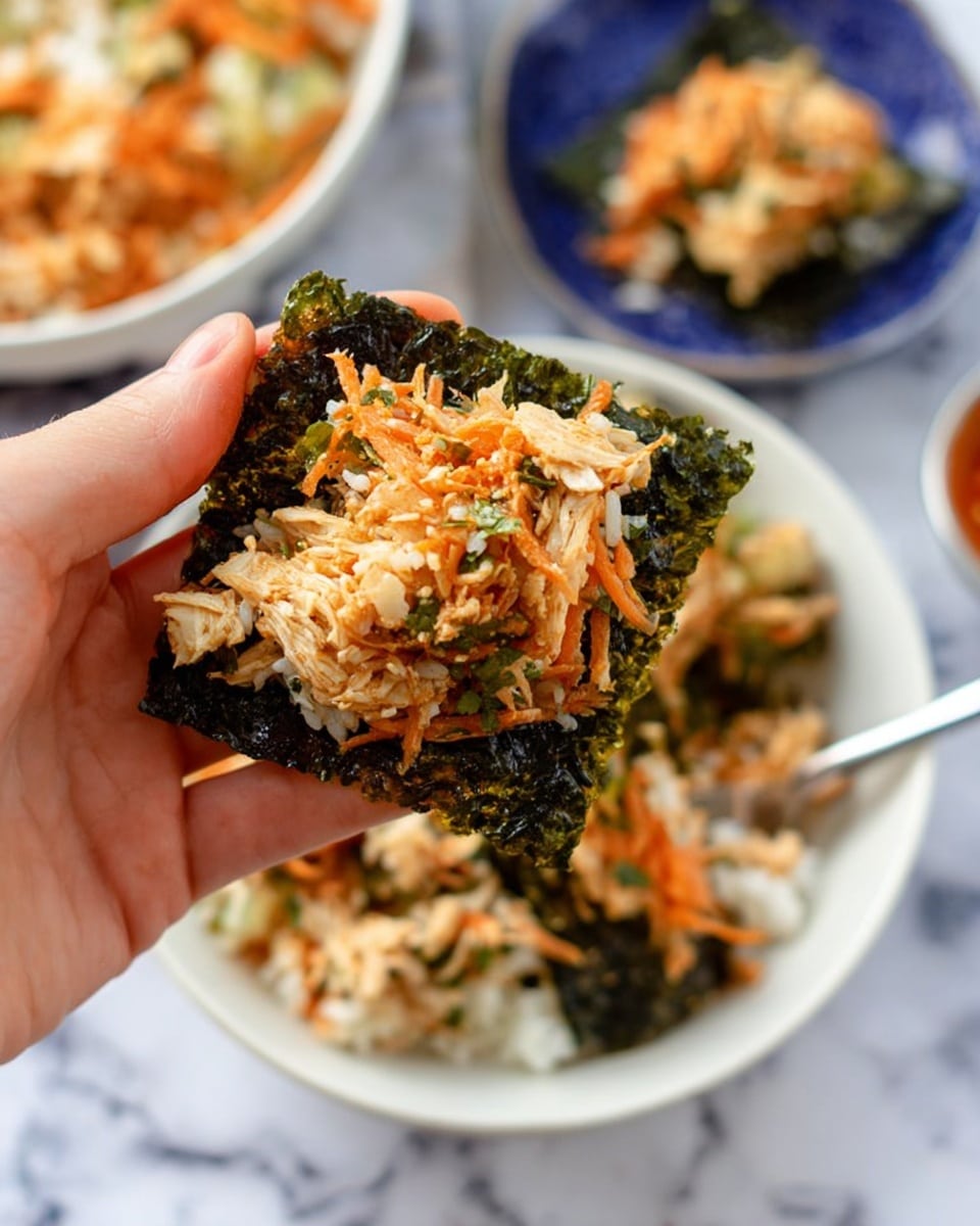 A woman's hand holds a dark green, crispy seaweed square topped with shredded orange carrot, light tan shredded chicken, and bits of white rice mixed with small green herbs. Behind this, in a white bowl with a spoon, there is a mix of white rice, shredded chicken, and some green vegetable pieces. The bowl sits on a surface with a white marbled texture, and blurred food items are in the background. The colors show a fresh mix of greens, oranges, tans, and whites, giving a layered look of seaweed, vegetables, and rice. photo taken with an iphone --ar 4:5 --v 7