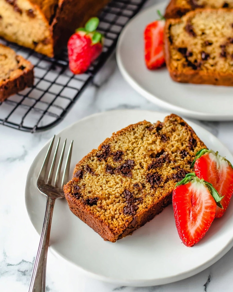 The image shows one slice of golden brown chocolate chip bread with visible small dark chocolate chips spread throughout the soft and slightly crumbly texture, placed on a white plate on the lower part of the image. Next to the bread slice, there are three whole bright red strawberries with green leaves and one strawberry sliced in half showing its juicy, red inside. A silver fork is placed to the left of the bread slice on the plate. In the background, another white plate partially visible holds another slice of the same chocolate chip bread, and more slices of bread rest on a black cooling rack on a white marbled surface. Photo taken with an iphone --ar 4:5 --v 7