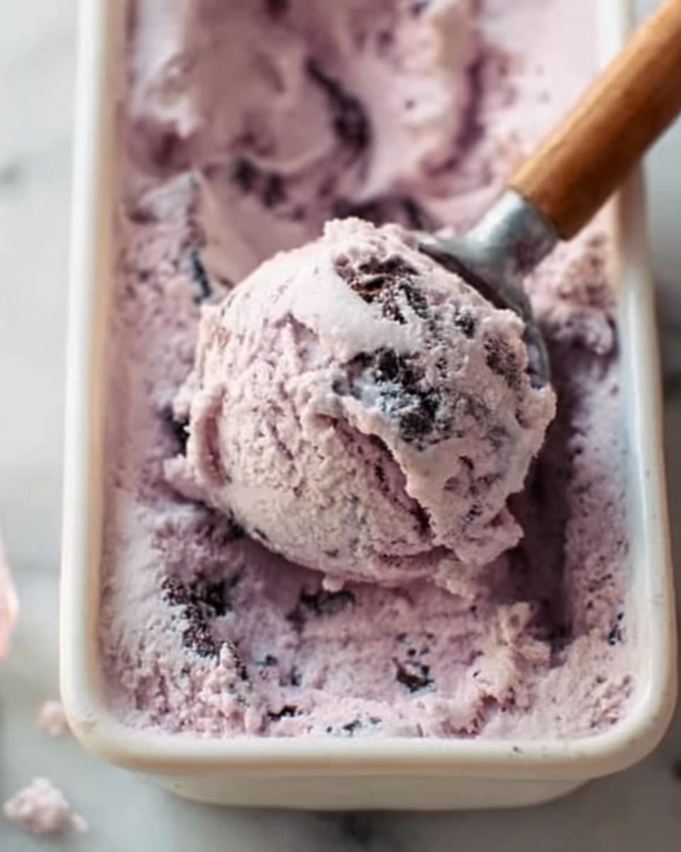 The image shows a close-up of a scoop of light purple ice cream with visible dark chunks inside it, placed in a rectangular white container. A spoon with a wooden handle is inserted into the ice cream, and the texture looks creamy and soft with some rough areas where the scoop was taken. The background is a white marbled surface, giving the scene a clean and fresh feel. photo taken with an iphone --ar 4:5 --v 7