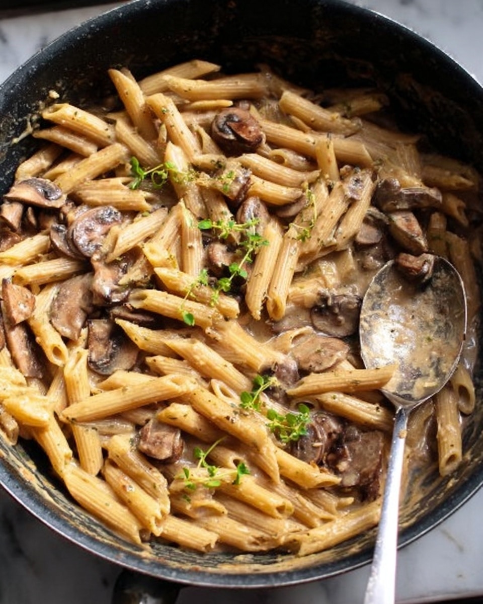 The image shows pasta penne cooked with mushroom slices and small green herb leaves, all mixed in a creamy light brown sauce inside a black pan. A large silver spoon is resting in the pan, partly covered with the same sauce. The pasta pieces are light beige and lightly shiny from the sauce. The mushroom slices are dark brown and soft looking, well spread among the pasta. The small green herb leaves add a touch of fresh color on top. The pan has some sauce splattered on the sides. The whole scene is on a white marbled surface. photo taken with an iphone --ar 4:5 --v 7