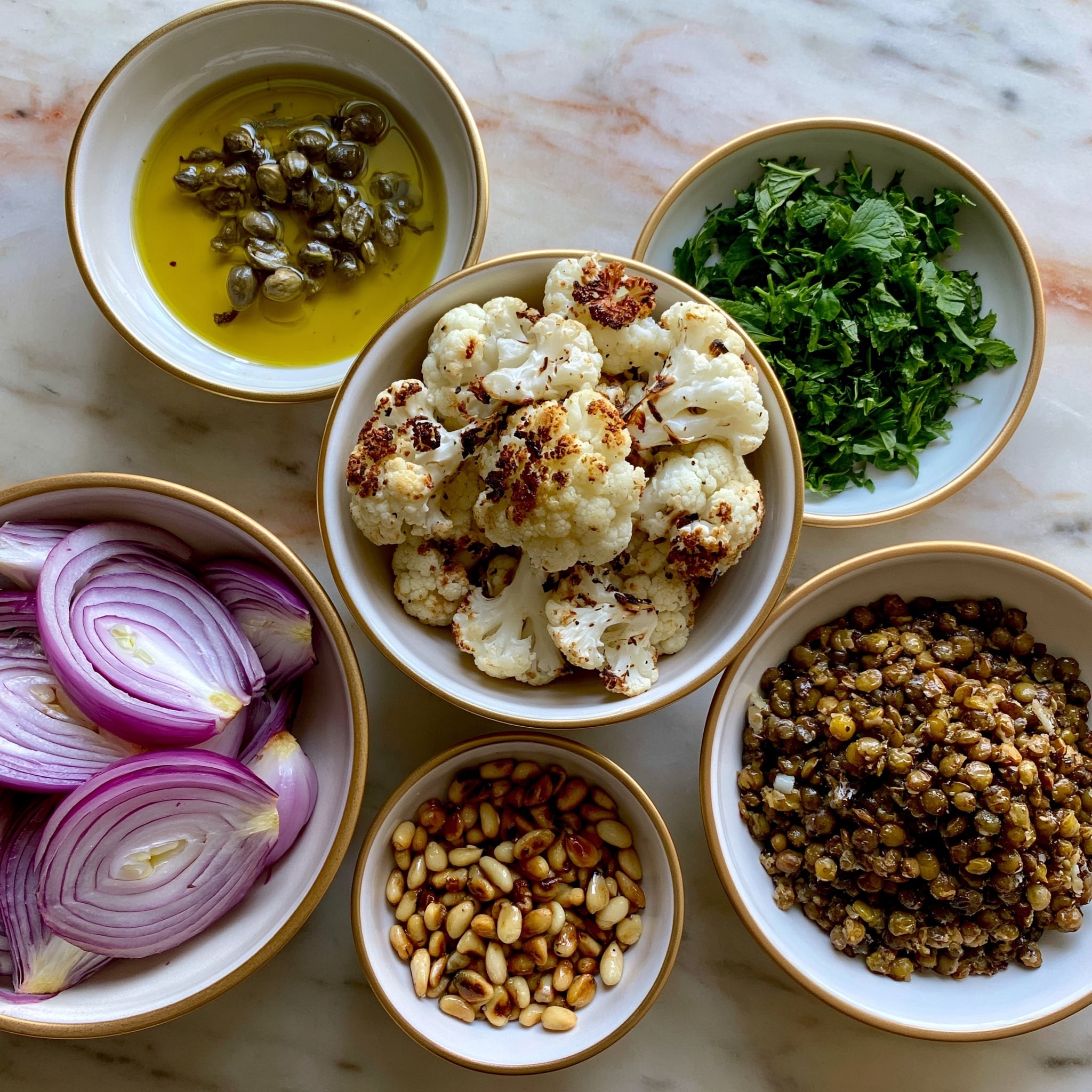 A large white bowl filled with cooked cauliflower florets that are light golden brown on the edges, mixed with small round lentils and thin slices of mushrooms. The dish is garnished with small pieces of fresh green herbs scattered throughout. A silver spoon and a wooden spoon rest inside the bowl on the right side. Around the bowl on a white marbled surface, there are some loose pine nuts and parsley leaves, and next to it, a small white plate holds finely chopped green herbs. A white cloth is positioned at the top left corner of the image. Photo taken with an iphone --ar 4:5 --v 7