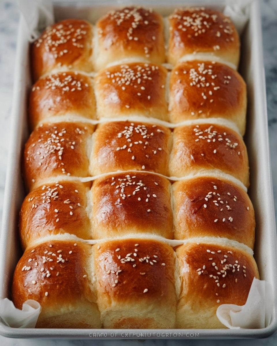 The image shows a white baking tray filled with two rows of soft, golden brown bread rolls, each roll topped with small white sesame seeds. The rolls have a glossy surface, a fine crust, and are arranged side by side, separated by thin white lines of dough on top, creating a neat, connected pattern. The bread looks fluffy and slightly puffy, with smooth, shiny tops and light brown edges. The tray is lined with white parchment paper, and the background is a white marbled texture. photo taken with an iphone --ar 4:5 --v 7