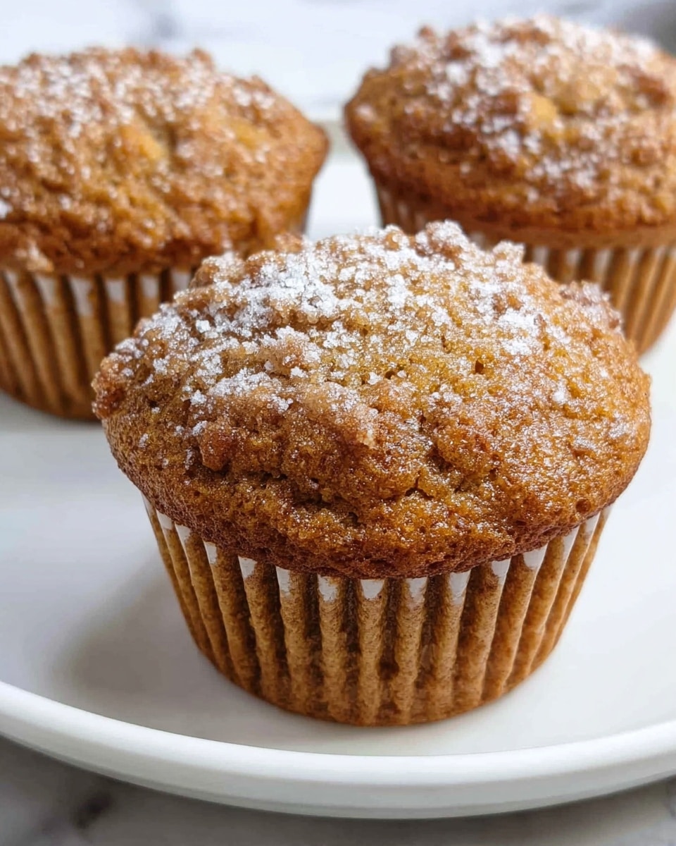 The image shows three golden brown muffins on a white plate, each with a slightly rough and crumbly texture on top. The muffins have a light dusting of white powdered sugar sprinkled evenly over their tops. The muffin wrappers are a light brown color with vertical ridges, tightly holding the muffins. The surface beneath the plate has a white marbled texture. The focus is on the muffin in front, with the other two muffins softly blurred in the background. photo taken with an iphone --ar 4:5 --v 7
