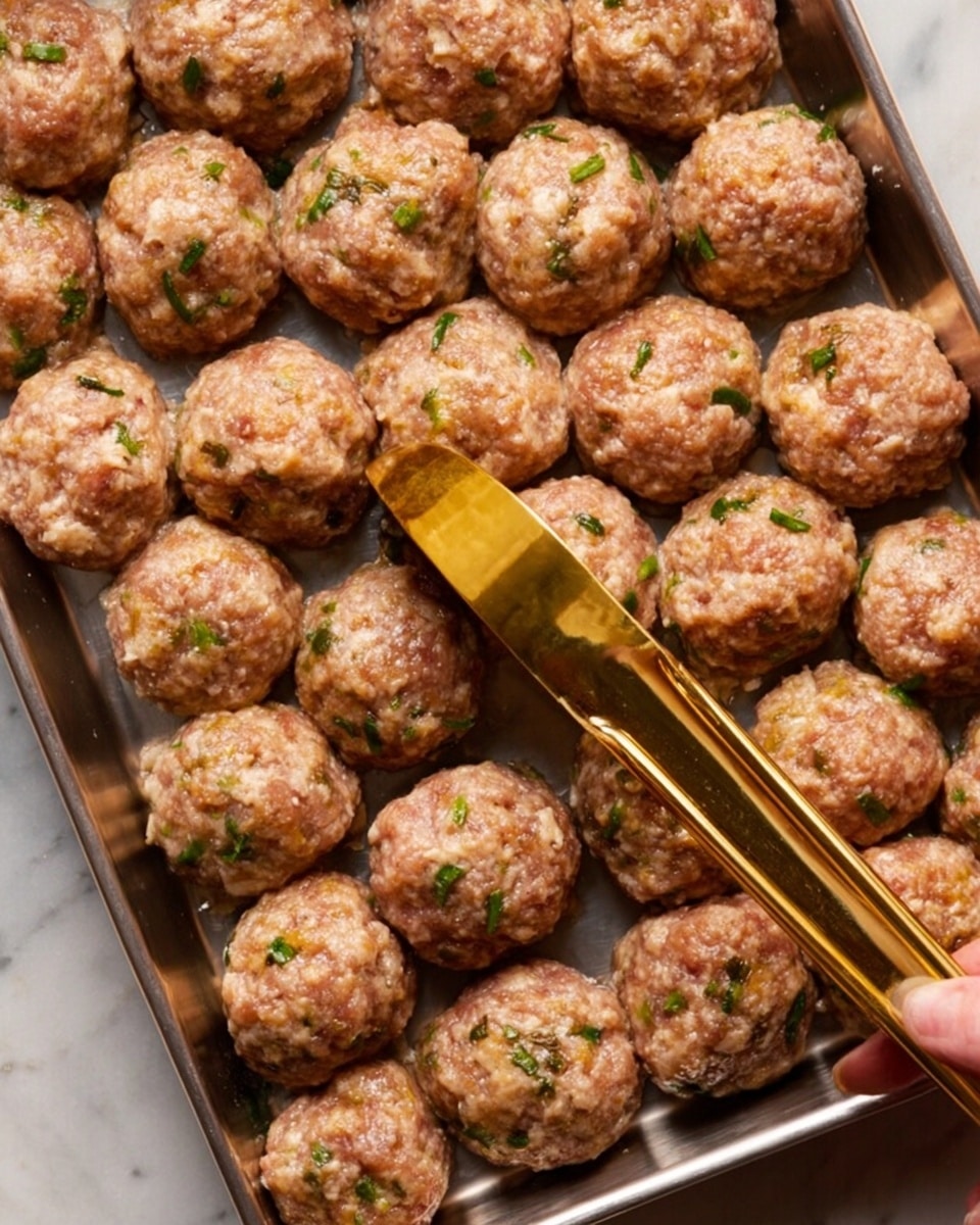 The image shows a metal tray filled with evenly shaped raw meatballs arranged closely in neat rows. Each meatball is rough-textured with visible pieces of green herbs mixed in, giving small specks of color among the light brown meat. A shiny gold-colored scoop is placed diagonally on top of the meatballs, with a woman's hand holding the handle, suggesting the meatballs were shaped using this tool. The tray rests on a white marbled surface. Photo taken with an iphone --ar 4:5 --v 7