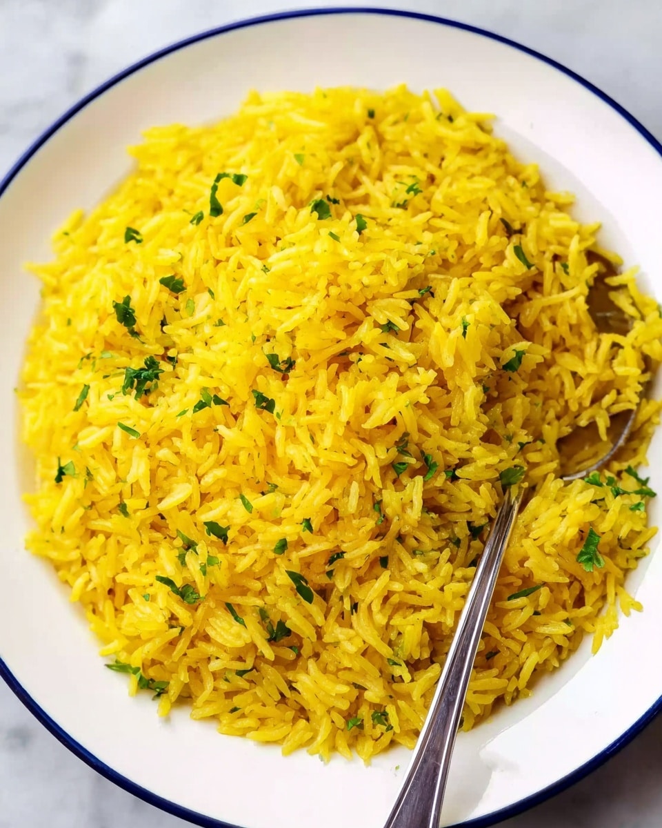 A close-up top view of a white plate filled with bright yellow rice mixed with small green herb pieces. The rice grains are long and fluffy, showing a soft texture. A silver spoon is partially buried in the rice, with its handle resting on the plate's edge. The white plate has a thin blue rim and is placed on a white marbled surface. photo taken with an iphone --ar 4:5 --v 7