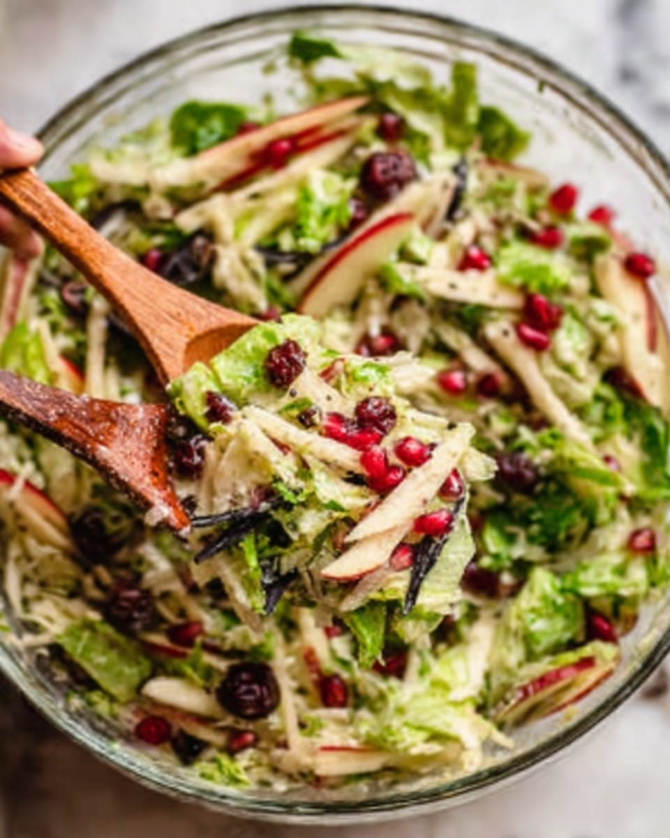 A clear glass bowl filled with a fresh salad shows different layers: pale green lettuce leaves create the base, mixed with lighter slices of apple and red pomegranate seeds sprinkled on top. There are small black olives and thin black strips that add texture. Two wooden spoons rest inside the bowl, gently lifting the salad, with a woman's hand holding one of the spoons. The background surface is white marble. photo taken with an iphone --ar 4:5 --v 7