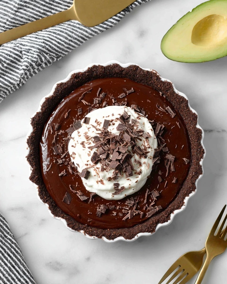 The image shows a chocolate tart on a white marbled surface. The tart has three clear layers: a dark brown crust on the bottom with a rough texture, a smooth, glossy dark chocolate filling in the middle, and a dollop of white whipped cream in the center on top. The whipped cream is sprinkled with uneven dark chocolate shavings that spread lightly on the tart's surface around the cream. The tart is in a fluted white tart pan. To the right of the tart, there is half a bright green avocado with its seed removed. Near the bottom left corner are gold-colored fork and knife placed next to each other. On the top right corner, there is a folded black and white striped cloth. At the top left, a gold-colored cake server rests on the white marbled surface. The photo taken with an iphone --ar 4:5 --v 7