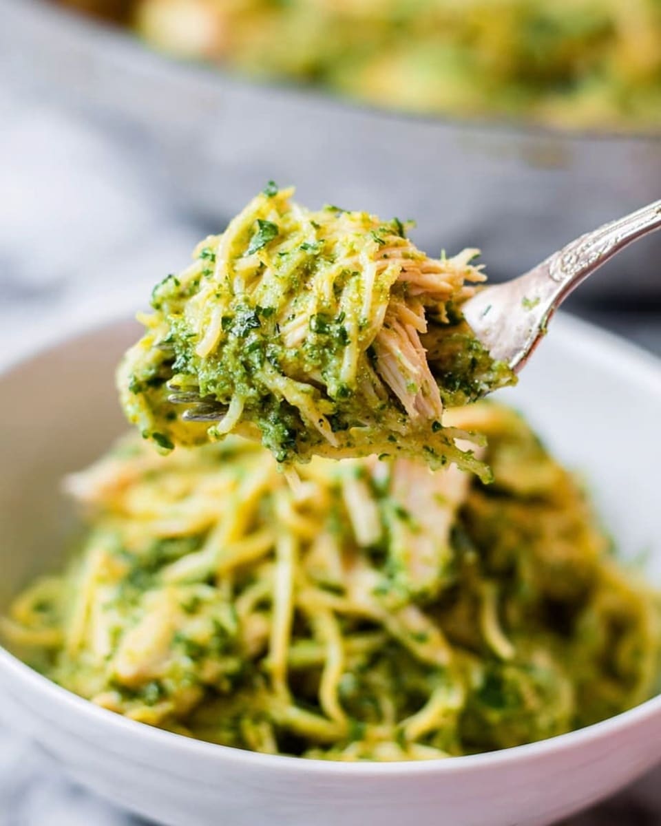 A close-up view of a fork lifting a small bunch of shredded pasta mixed with thick green pesto sauce and pieces of shredded light-colored chicken from a white bowl. The pasta strands are thin and coated evenly with the bright green sauce that has small bits of herbs, giving it a textured look. In the blurred background, there is a larger white bowl also filled with the same green pasta. The scene is set on a white marbled surface. Photo taken with an iphone --ar 4:5 --v 7
