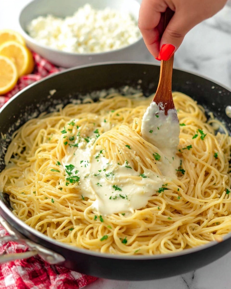 A close-up of a black pan filled with cooked spaghetti noodles in the center, light yellow in color with a smooth texture. Creamy white sauce is being stirred into the noodles from the left side of the pan with a wooden spoon held by a woman's hand with red nail polish. Small green parsley pieces are sprinkled on top of the noodles and sauce. In the blurred background, there is a white bowl of cottage cheese on a red and white checkered cloth, and some lemon slices on a white marbled surface. Photo taken with an iphone --ar 4:5 --v 7