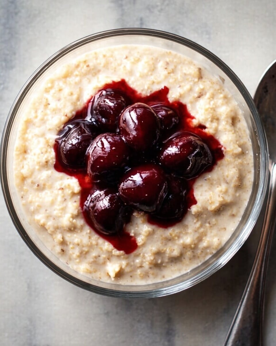 In a clear round glass bowl, there is a creamy light beige porridge with a soft texture, filling almost the entire bowl. On top, near the center, there are six dark red cherries with a shiny and wet look, slightly bursting in places, creating small pools of deep red juice that contrast with the lighter base. The bowl sits on a white marbled surface with a silver spoon placed to the right side. The scene is bright and simple, focusing on the colors and textures of the food. Photo taken with an iphone --ar 4:5 --v 7