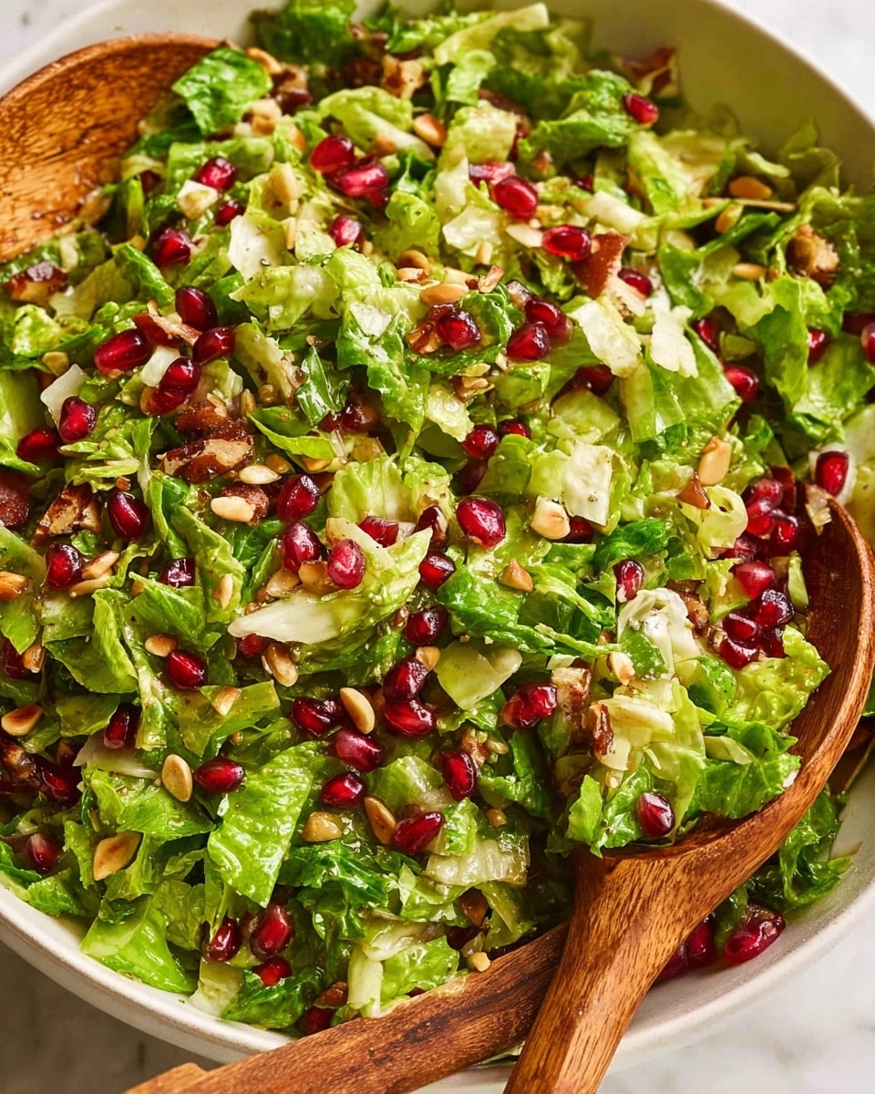 A close-up view of a fresh salad in a white bowl on a white marbled surface, showing finely chopped bright green lettuce leaves scattered all around. Among the lettuce pieces are shiny, deep red pomegranate seeds and small, light brown pine nuts evenly spread in the mix. There are also thin slices of pale beige nuts or seeds adding texture. The salad is being lightly mixed with two wooden salad spoons placed inside the bowl, adding a warm brown contrast to the vivid greens and reds. photo taken with an iphone --ar 4:5 --v 7