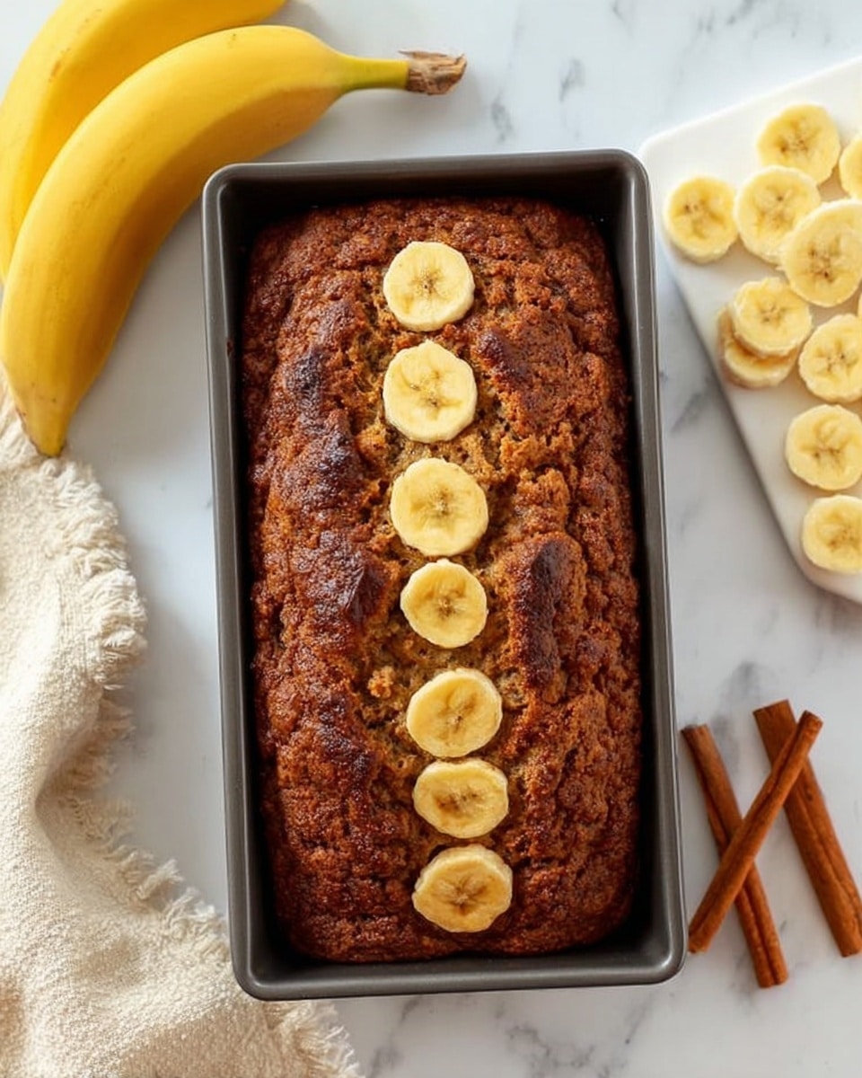 A loaf of banana bread sits in a dark metal pan on a white marbled surface, with seven round slices of banana arranged in a vertical line down the center of the bread's brown, textured top. To the left, there are two whole yellow bananas, and to the right is a small white wooden board with many round banana slices neatly laid out on it. Near the bottom right corner, two cinnamon sticks lie on the white marbled surface. A cream-colored cloth is partly visible in the bottom left corner. Photo taken with an iphone --ar 4:5 --v 7