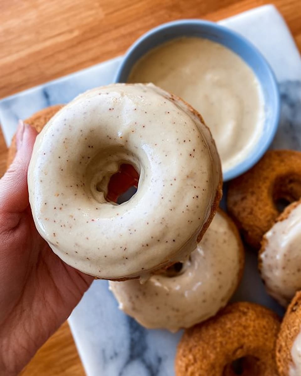 A close-up of a woman's hand holding a single donut that is fully covered with a smooth, light beige icing with tiny specks, showing a creamy and shiny texture. Below the donut, there is a white bowl filled with the same beige icing. Around the bowl on a white marbled surface, there are several donuts, some plain and some partially covered with icing. The background is a light wooden surface. Photo taken with an iphone --ar 4:5 --v 7
