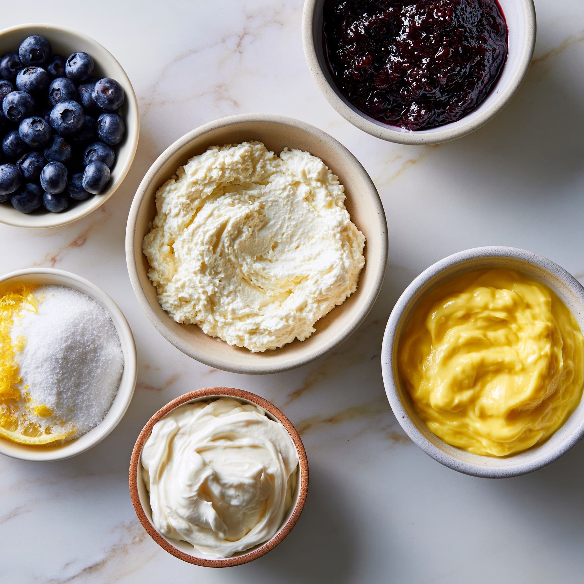 A large clear glass bowl holds a layered dessert with five visible layers starting with a pale yellow creamy base, followed by small golden cake pieces and fresh dark blue blueberries repeated several times. The top layer is covered with more blueberries and five dollops of white whipped cream arranged evenly around the edge. Each whipped cream swirl is topped with a small wedge of bright yellow lemon and one blueberry, two of the swirls have small green mint leaves on top as well. The bowl is set on a white marbled surface with bright yellow lemons in a white bucket in the background, and a small jar of yellow sauce and half a lemon nearby, with some purple flowers adding a splash of color. photo taken with an iphone --ar 4:5 --v 7