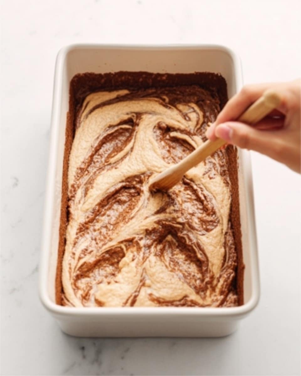A white rectangular baking dish filled with light brown batter that has swirls of darker brown mixed inside. A woman's hand is holding a small wooden stick, drawing circular patterns across the surface of the batter. The dish is set on a white marbled background. photo taken with an iphone --ar 4:5 --v 7