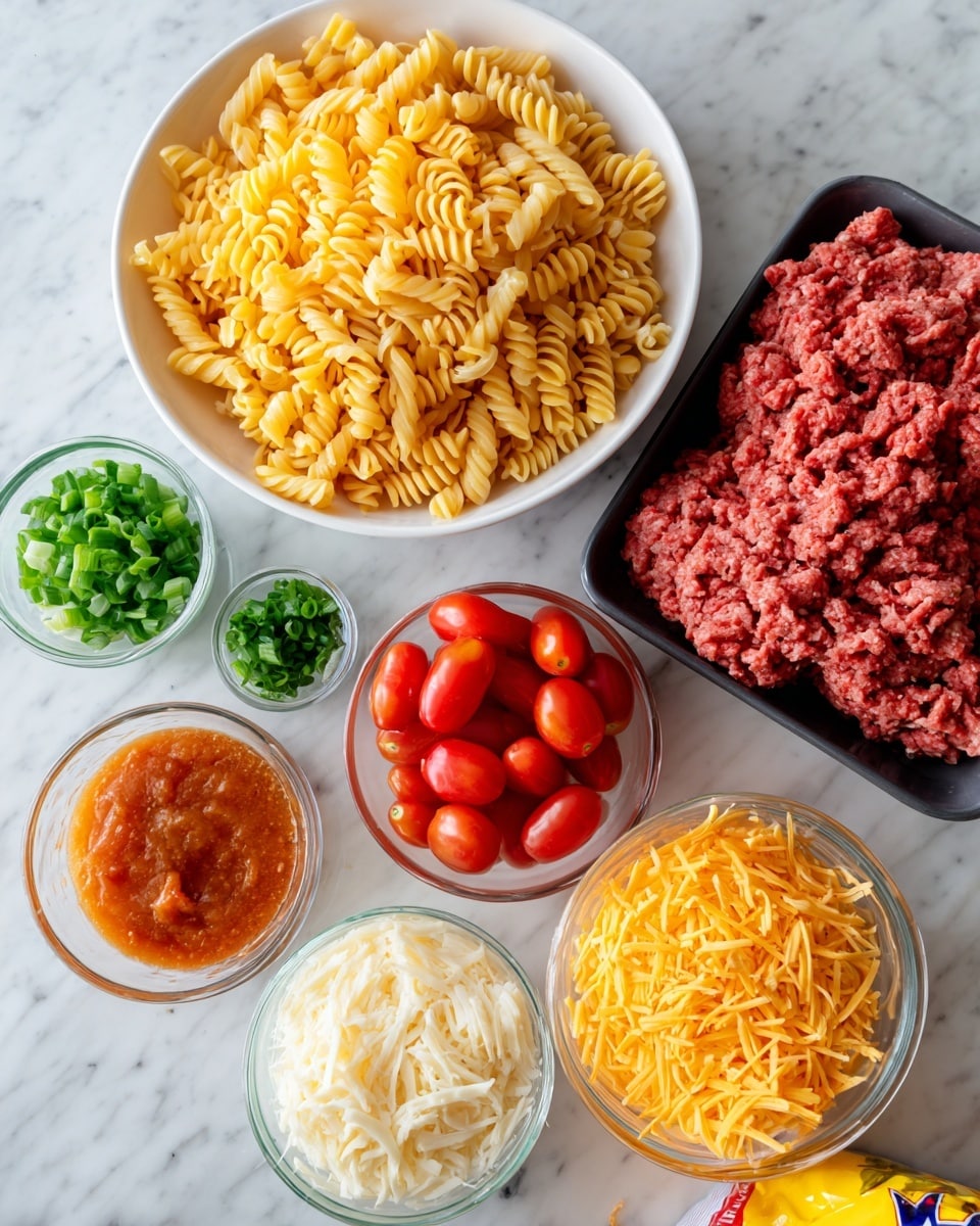 The image shows various ingredients arranged neatly on a white marbled surface. At the center bottom is a large, unopened red bag of Nacho cheese Doritos with visible chips inside. Above the bag, a white bowl filled with uncooked golden yellow rotini pasta sits to the left, while to the right is a black tray holding bright red raw ground beef. To the far right, there is a pile of halved cherry tomatoes with shiny red skins and soft interiors visible. On the bottom left, there are two small clear bowls, one with chopped green onions and the other with chopped green cilantro leaves. Just above these bowls is a glass container filled with reddish-orange Catalina dressing. Below the Doritos bag, two small piles of shredded cheese sit side by side on the white marbled surface—one is creamy white Monterey Jack cheese, and the other is rich orange cheddar cheese. To the bottom right of the Doritos bag is a yellow packet of taco seasoning mix labeled