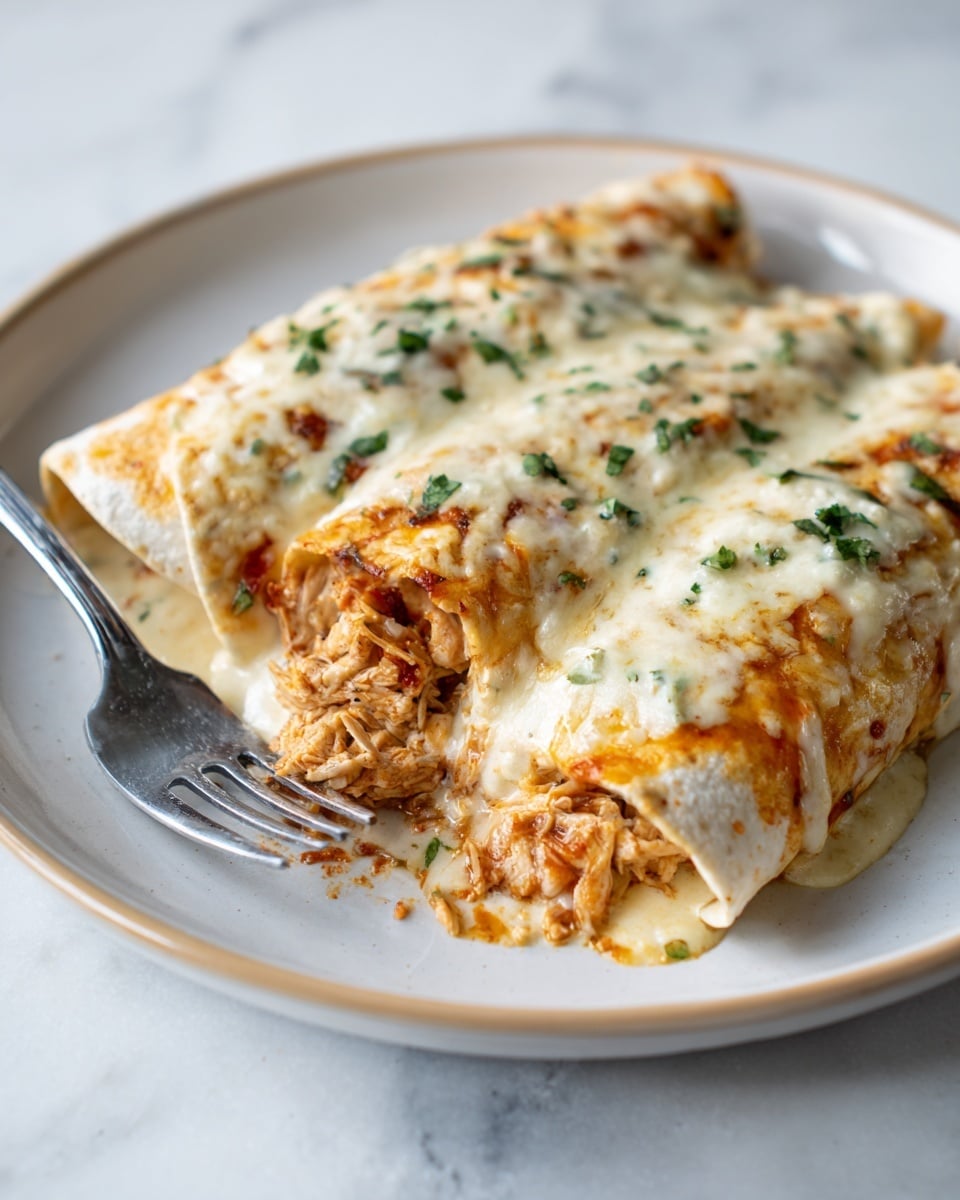 Two enchiladas lie side by side on a white plate with a subtle rim. Each enchilada is covered with a creamy, white cheese sauce topped with small green herb pieces. One enchilada is cut open, showing shredded, light brown chicken mixed with a red sauce inside. A silver fork rests on the front left side of the plate. The plate sits on a white marbled surface. photo taken with an iphone --ar 4:5 --v 7