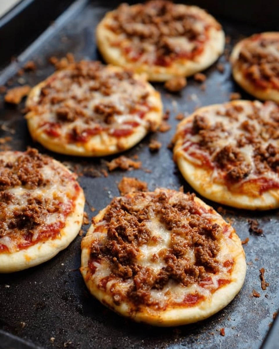 The image shows six small round pizzas placed on a dark baking tray with a slightly worn surface. Each pizza has a light golden crust, a visible thin layer of red tomato sauce just beneath a layer of melted, slightly browned cheese, and is topped with a generous, uneven crumbly layer of cooked ground meat. Around the pizzas, some bits of cooked meat and cheese are scattered on the tray. The focus is on the pizzas, with a shallow depth of field that blurs parts of the tray edges. Photo taken with an iphone --ar 4:5 --v 7