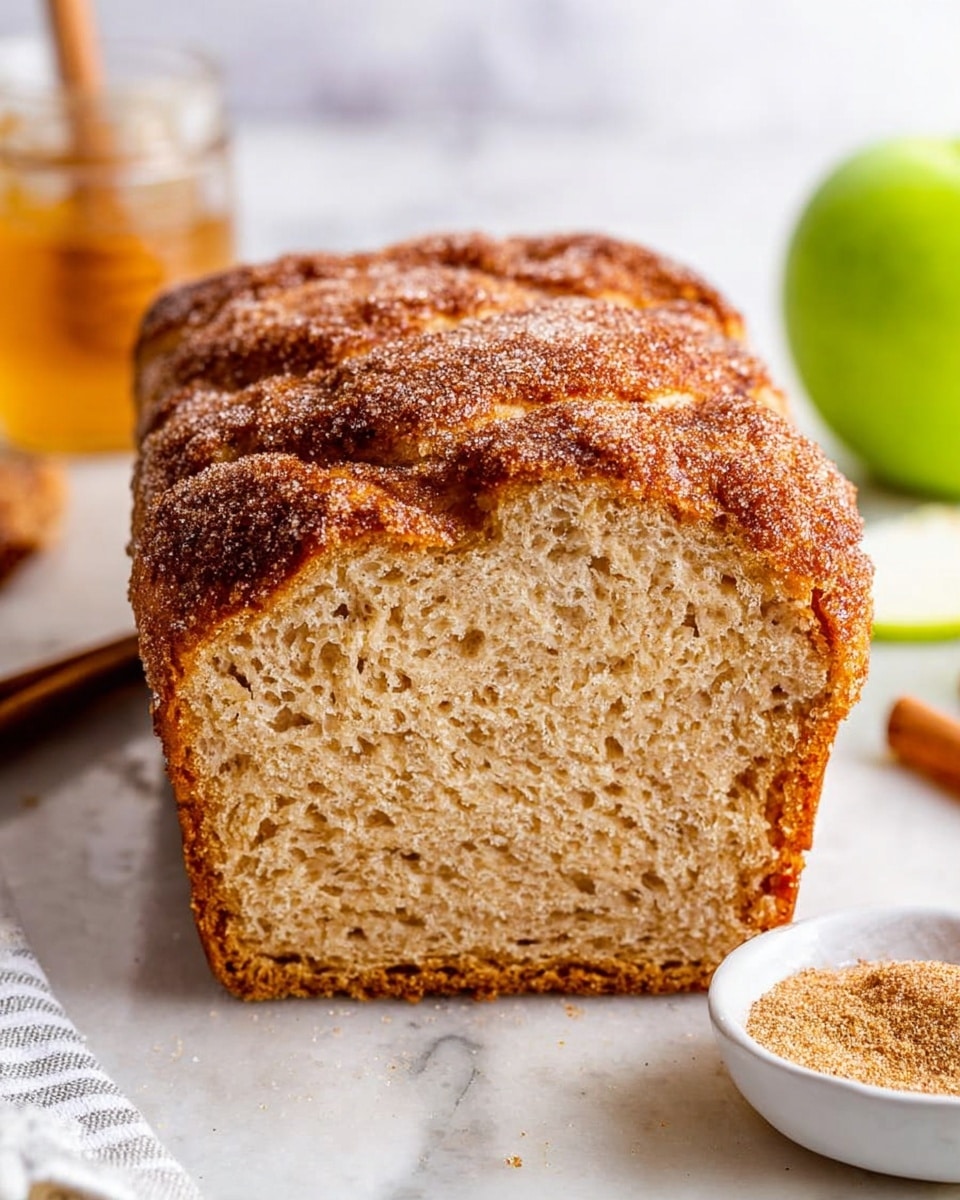 A thick slice of cinnamon bread is placed upright on a white marbled surface, showing a soft, light brown, moist inside with a slightly darker crust. The top of the bread is dusted with a layer of cinnamon sugar, creating a textured, crystalline look. In the background, there is a blurred green apple on the right side and a honey jar on the left side. The crust's ridged pattern rises across the top, giving a rustic feel. A small white bowl with a cinnamon sugar mix sits in the lower right corner, and a white cloth with subtle stripes is partly visible in the lower left. Photo taken with an iphone --ar 4:5 --v 7