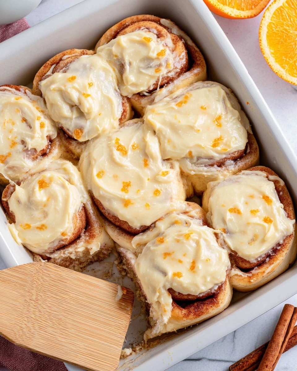 The image shows a white baking dish with eight cinnamon rolls arranged close together in two rows. Each roll has two layers: a light brown swirled base with a slightly golden baked texture and a thick layer of creamy pale yellow frosting spread unevenly on top with some small orange specks. The rolls look soft and moist, and one roll is slightly lifted on a light wooden spatula at the bottom left corner, showing the underside. The background is a white marbled texture with a half orange and a cinnamon stick on the right side. Photo taken with an iphone --ar 4:5 --v 7