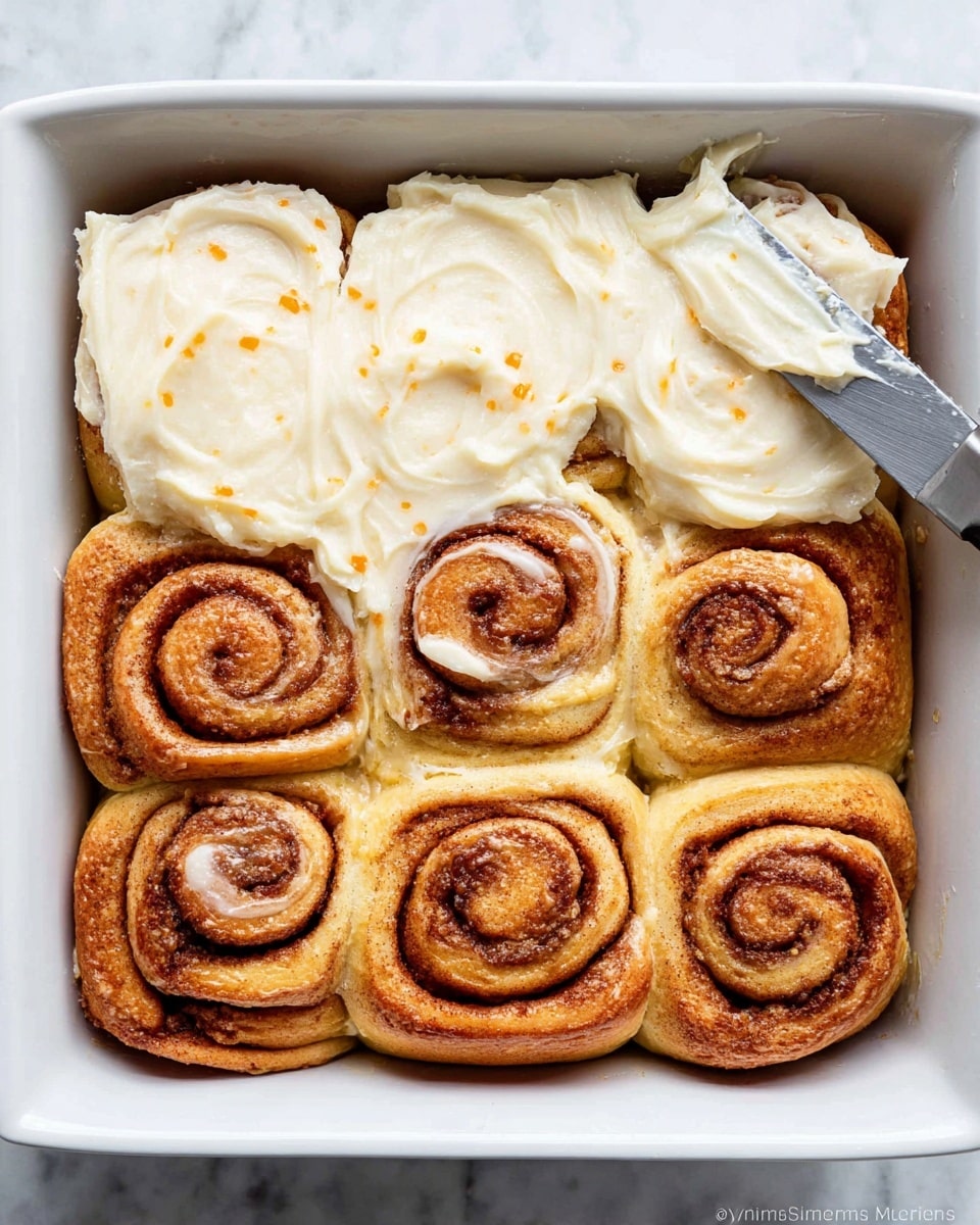 The image shows a white rectangular baking dish filled with nine cinnamon rolls arranged in a 3x3 grid. The bottom layer consists of golden-brown cinnamon rolls with visible swirls of cinnamon and sugar. The top layer is a thick, creamy white icing being spread evenly on the top six rolls with a metal butter knife, each roll partly covered with the smooth, shiny icing that has small orange specks. The top left three rolls remain uniced, showing their textured golden surface. The dish sits on a white marbled surface. photo taken with an iphone --ar 4:5 --v 7