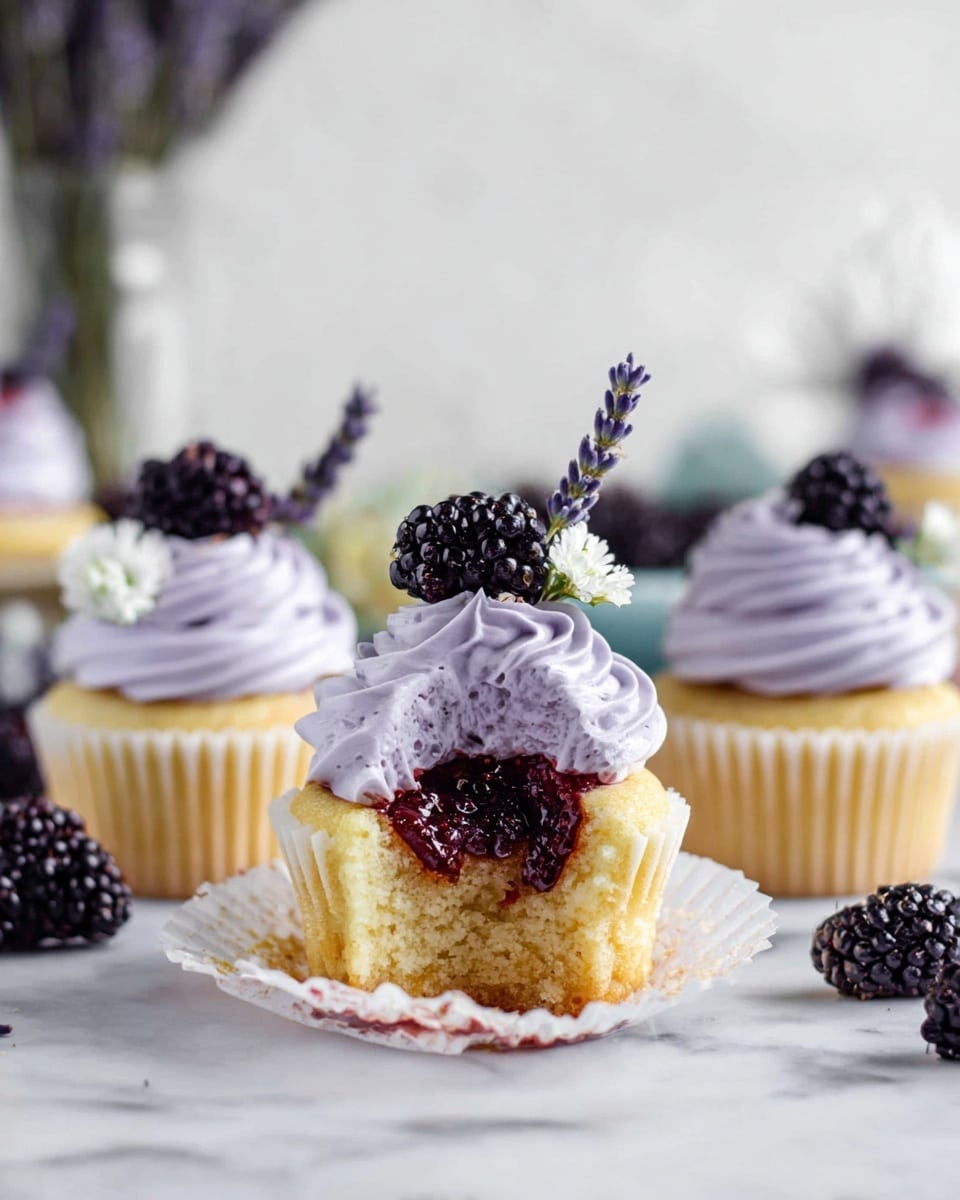 The image shows three cupcakes on a white marbled surface. Each cupcake has a golden yellow base with white wrappers, topped with light purple swirled frosting. On top of the frosting, there is a dark blackberry, a small white flower, and a sprig of purple lavender for decoration. One cupcake in the front is open, revealing a dark red jam center inside the soft yellow cake. The background is softly blurred, featuring more cupcakes, blackberries, a glass vase with lavender, and white elements. Photo taken with an iphone --ar 4:5 --v 7