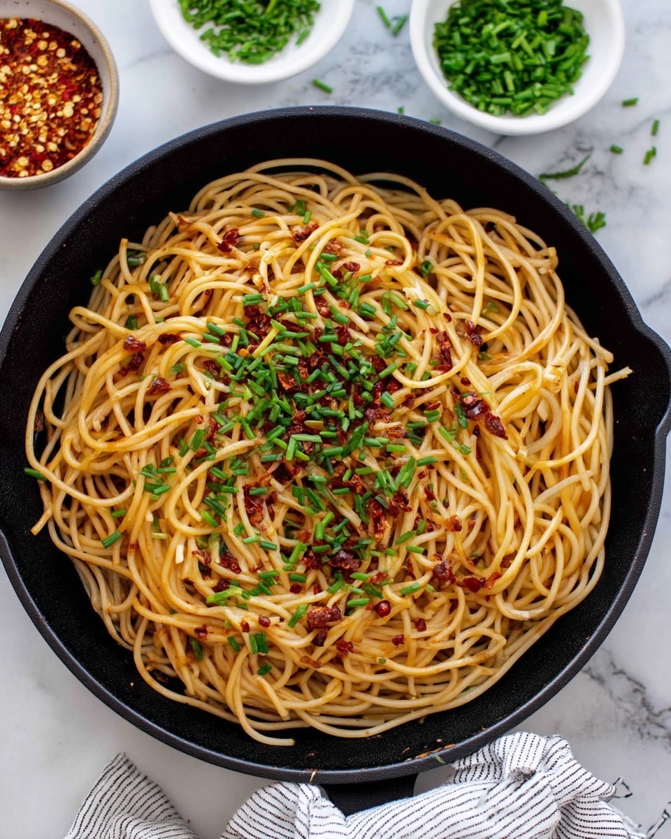 A black skillet filled with a single layer of light brown noodles, cooked and lightly tossed, covering the entire pan. The noodles are sprinkled with small pieces of reddish-brown chili flakes and bright green chopped chives scattered evenly on top. The skillet handle is wrapped with a white and gray striped cloth. There are two small white bowls placed near the skillet, one filled with red chili flakes and the other with chopped green chives. The background is a white marbled texture, with a few pieces of chives scattered around. photo taken with an iphone --ar 4:5 --v 7