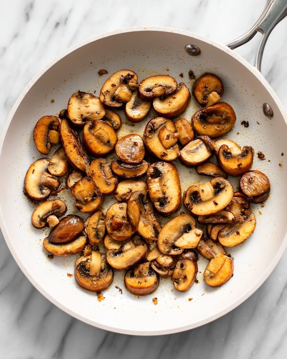 The image shows a white frying pan filled with cooked sliced mushrooms. The mushrooms are golden brown with a slightly crispy texture, spread evenly across the pan's surface. The pan sits on a white marbled surface, and some small browned bits are visible among the mushrooms, adding detail to their cooked appearance. Photo taken with an iphone --ar 4:5 --v 7